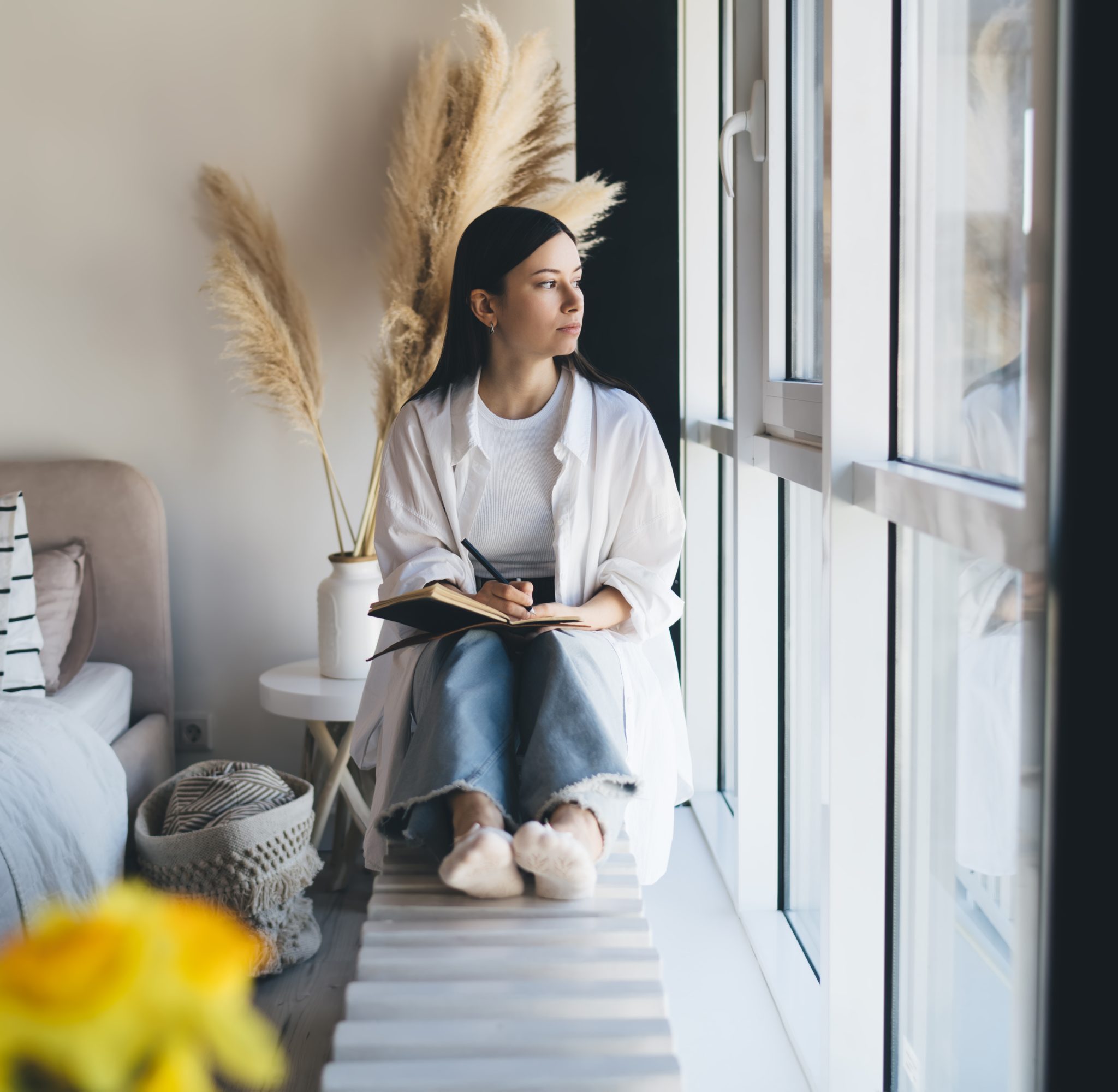 Thoughtful female in casual clothes taking notes in organizer and looking away while sitting on windowsill and chilling in modern bedroom